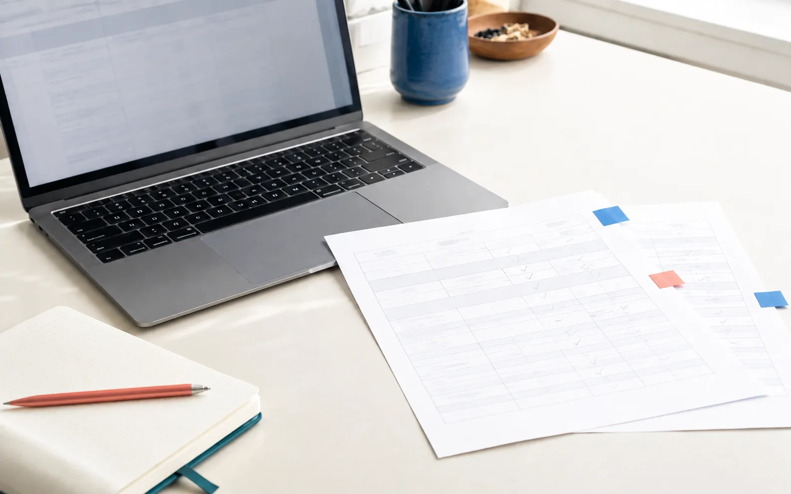 Bright comparison workspace with a laptop, white information sheets, colorful page tabs, and a blue pen cup.