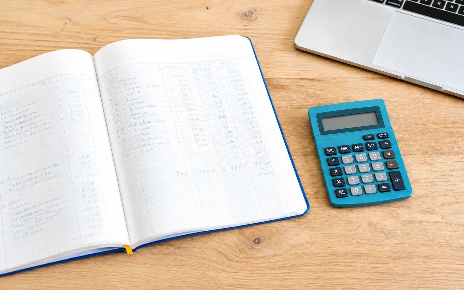 An open ledger with cobalt binding and a teal calculator on a sunlit wood desk.