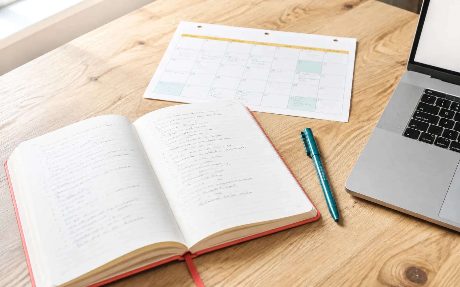 An open notebook with coral binding, teal pen and monthly calendar on a bright desk.