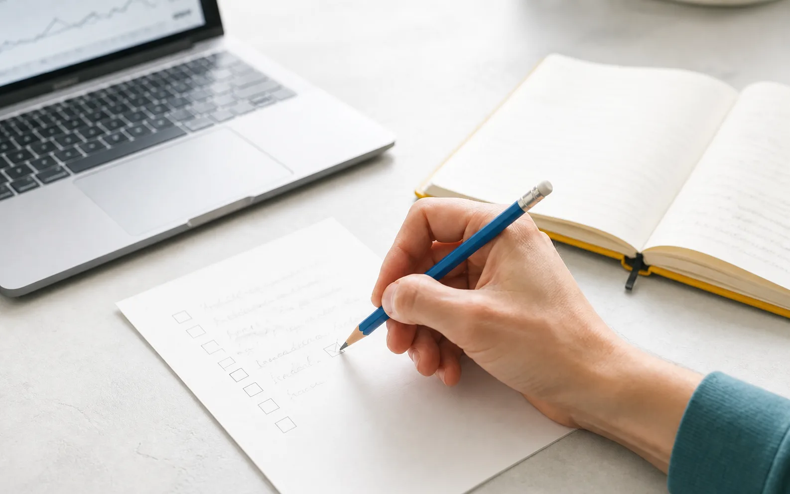 A hand checks a paper trading plan beside a laptop and mustard-edged notebook in bright daylight.